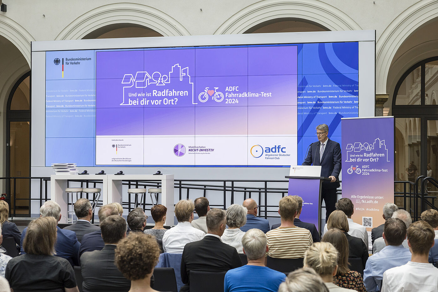 Mann mit grauen Haaren und Brille in dunklem Anzug steht am Rednerpult vor großer lila Leinwand mit "Und wie ist Radfahren bei dir vor Ort? ADFC Fahrradklima-Test 2024", Publikum sitzt in Reihen davor